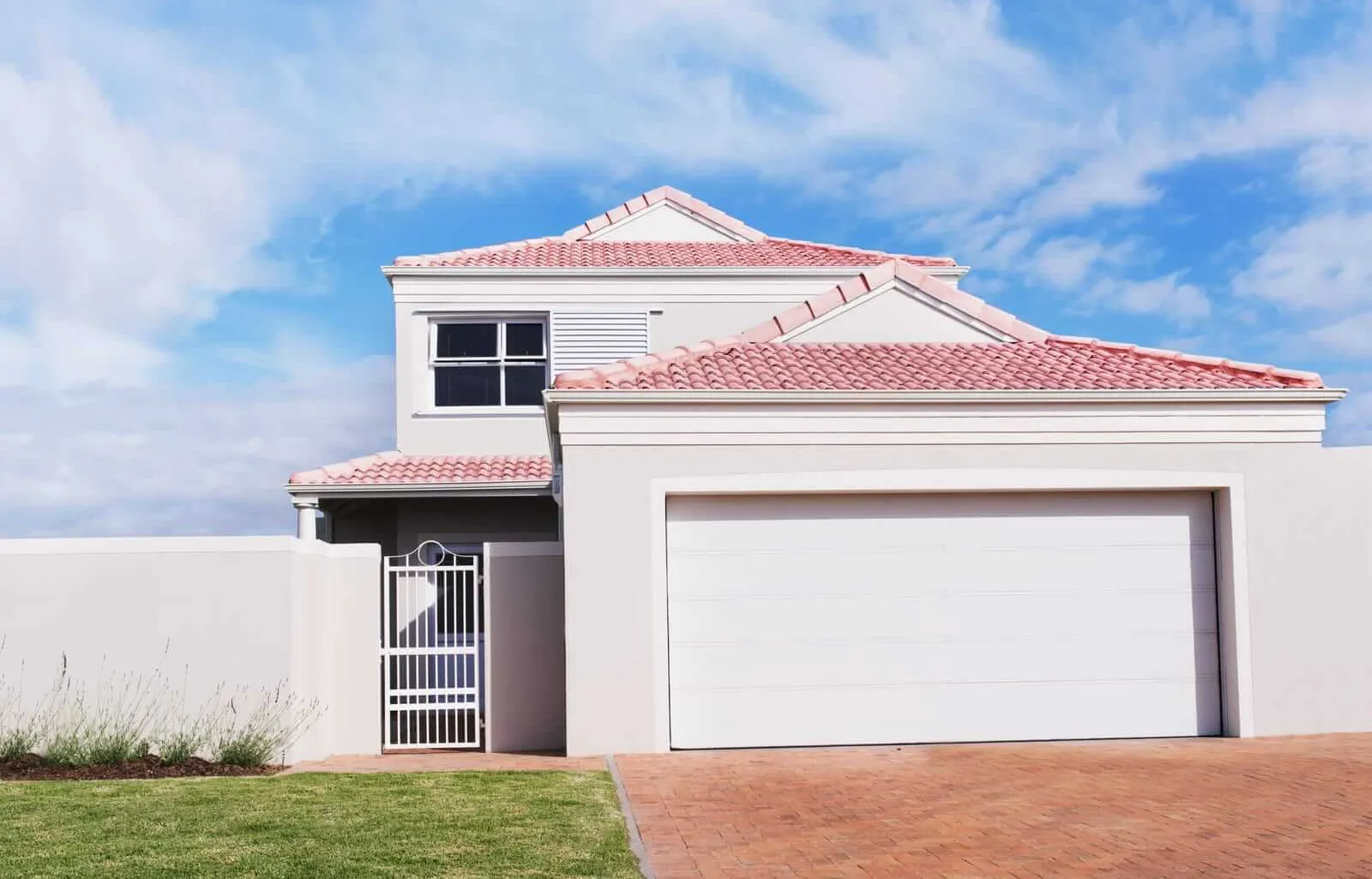 A modern two-story white house with red-tiled roofs and a double garage. The entrance features a metal gate, and the driveway is paved with red bricks. A low wall surrounds the property, with a small patch of grass in the foreground.