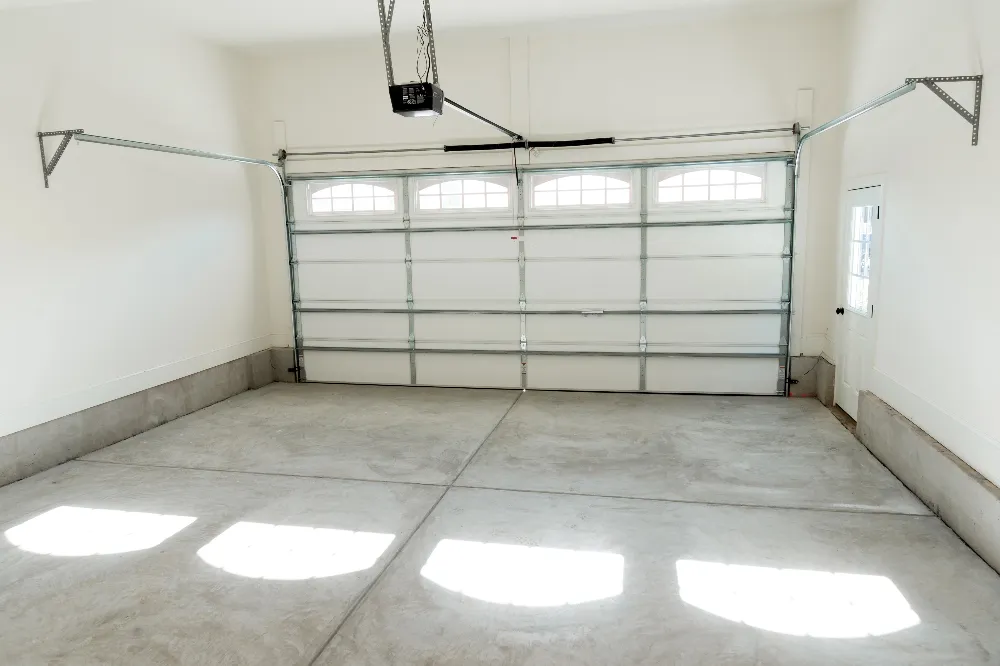 Empty residential garage with a closed white sectional door featuring four windows. The space has a smooth concrete floor and white walls. A motorized garage door opener is visible on the ceiling. Natural light shines through the windows.