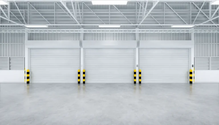 An empty industrial warehouse interior with three large closed garage doors. The space is well-lit with bright overhead lights, and the floor is smooth concrete. Yellow and black safety bollards stand in front of each door.