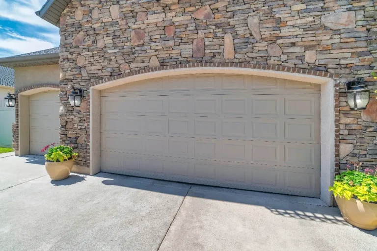 A stone-clad house features two adjacent beige garage doors. Each door is framed by outdoor lantern-style light fixtures. Potted plants with colorful flowers flank the garage doors on a sunlit concrete driveway.