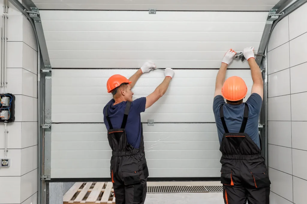 Two workers in orange helmets and black overalls focus on garage door maintenance. They face the door, carefully adjusting it with their hands, while a wooden pallet rests to the left.