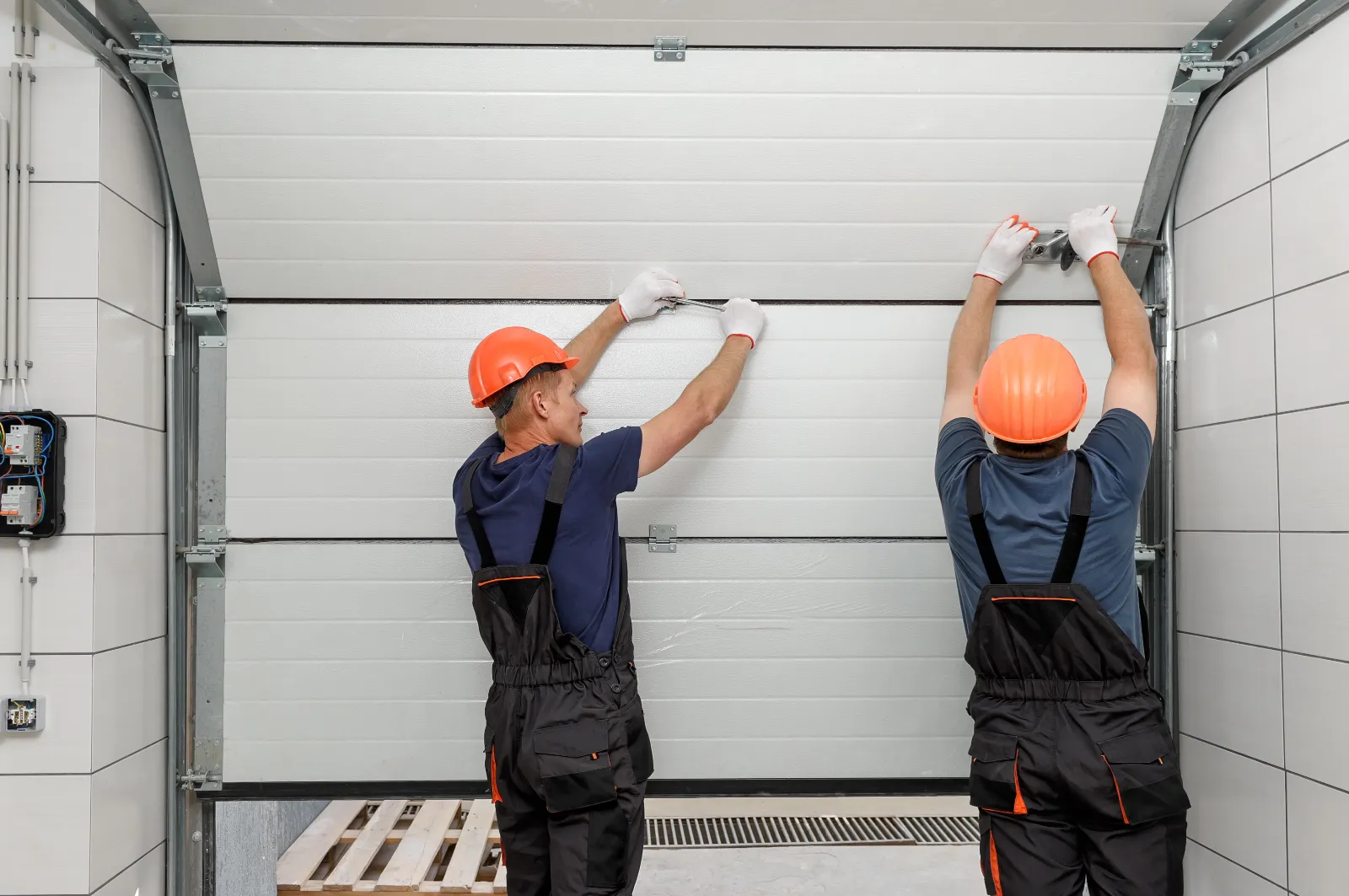 Two workers wearing orange helmets and black overalls are installing or repairing a garage door. They are adjusting the panels and working together inside a garage area.