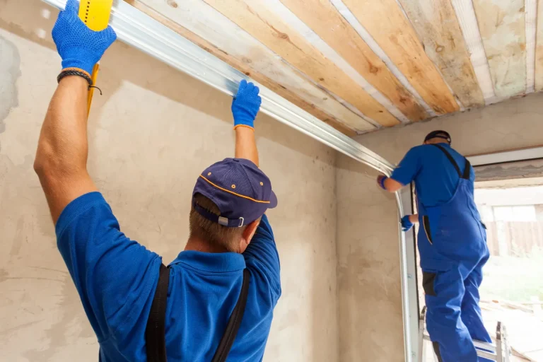 Two construction workers in blue uniforms and gloves are installing a metal beam under a wooden ceiling. One is on a ladder, while the other supports the beam from the ground. The room has unfinished walls.