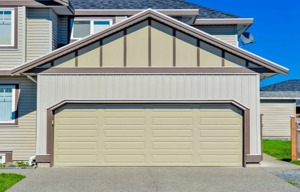 A beige house with a large garage door. The building features siding panels and a peaked roof. There is a concrete driveway leading to the garage, and the sky is clear and blue.