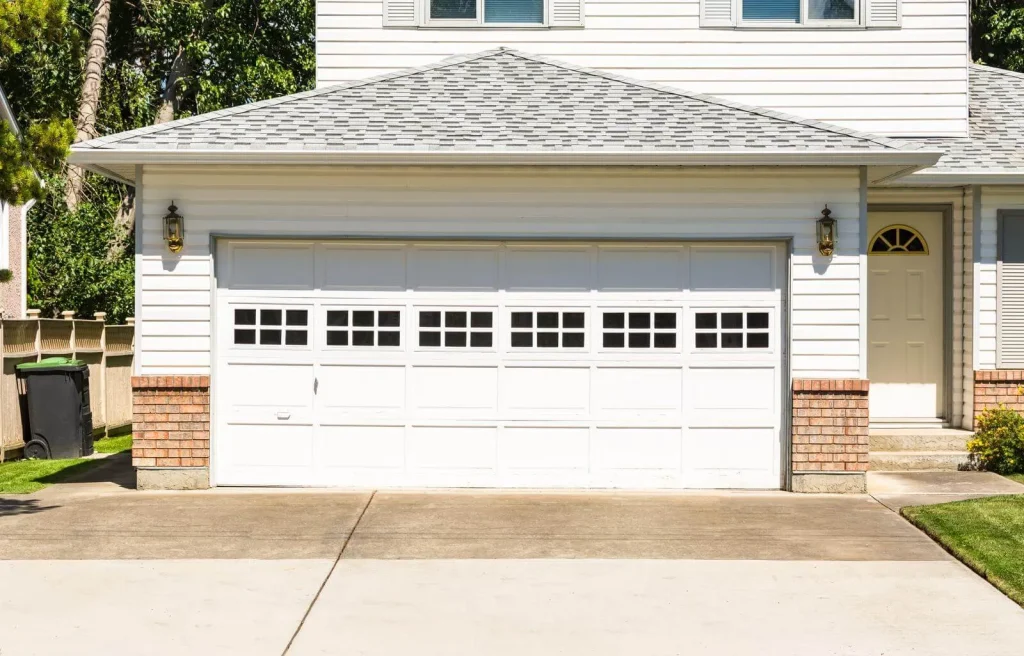 A suburban house with a white double garage door featuring square windows. The exterior is white with brick accents. There is a driveway leading up to the garage and a small lawn area with a trash bin on the side.