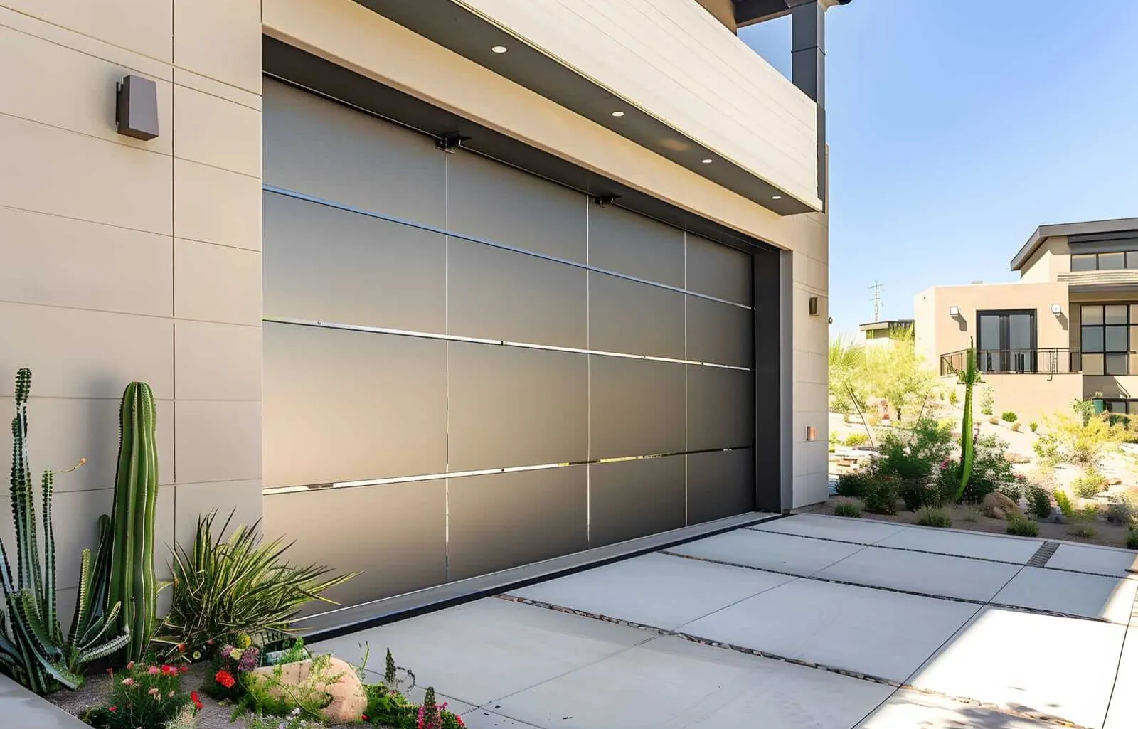 Modern garage with a sleek metallic door, surrounded by a minimalist landscape. The area features desert plants, including cacti, and a clean concrete driveway. Contemporary architecture is visible in the background.