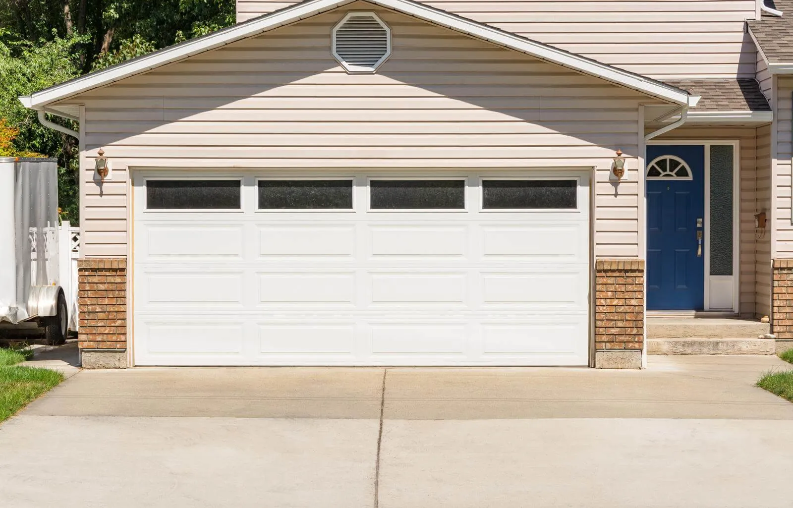 A two-car garage with a white door is attached to a beige house. The house features brick accents and a blue front door with an arched window. A light fixture is mounted near the garage, and a driveway leads up to it.