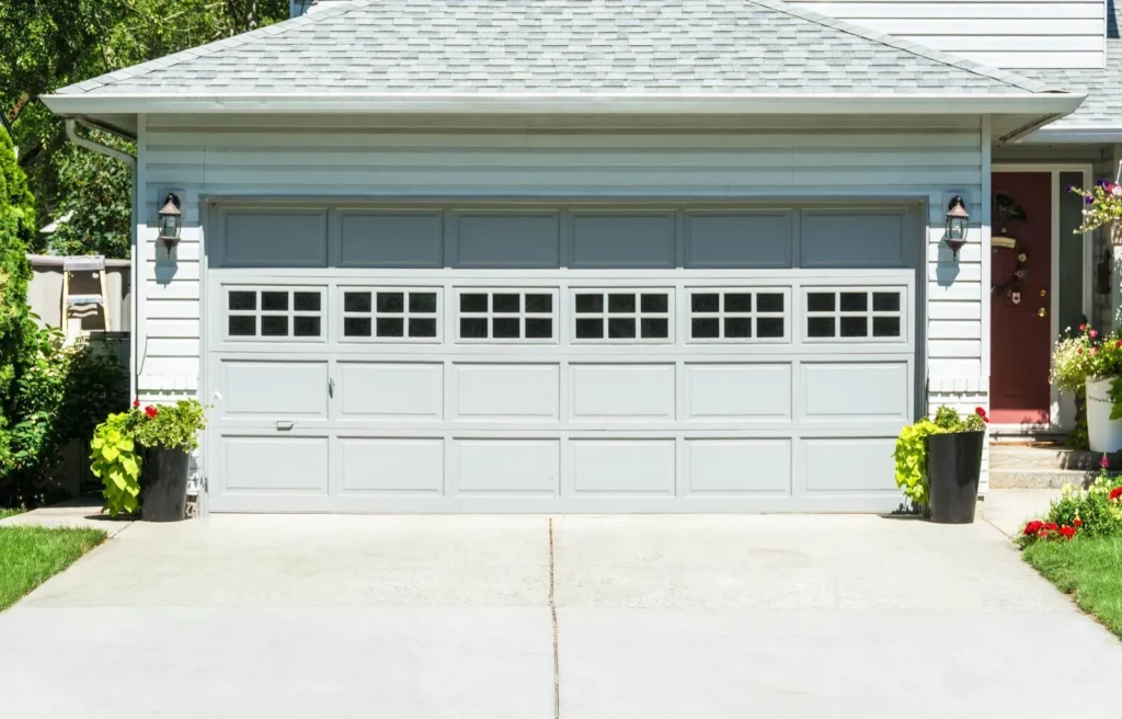 A gray garage door with windows in the top panels is centered on a white house. Two black planters with green and yellow foliage sit on either side. The driveway is concrete, and there are small shrubs around the edges.