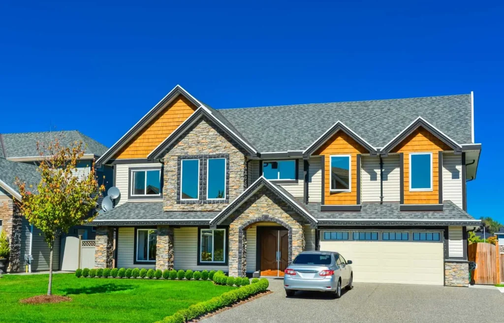 A modern two-story house with brick and wood accents, featuring large windows and a double garage. A silver car is parked in the driveway, and a well-manicured lawn with a small tree is in the foreground. Clear blue sky in the background.