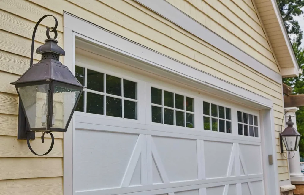 Exterior view of a yellow house with a white garage door featuring multiple glass panes at the top. Two black metal lantern-style lights are mounted on either side of the garage entrance. Trees are visible in the reflection of the garage windows.